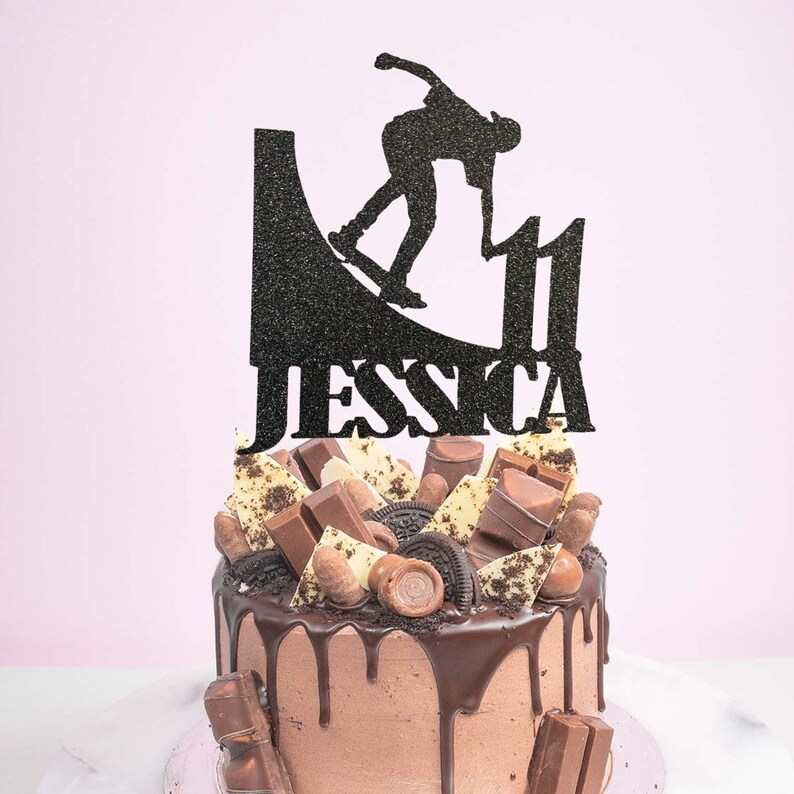 May include: Black glitter cake topper with a silhouette of a person skateboarding on a ramp. The topper says "Jessica" and the number "11".