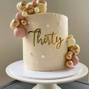 May include: A beige frosted cake with the word "Thirty" in gold script. The cake is decorated with gold, pink, and white sphere-shaped ornaments and sits on a white cake stand.