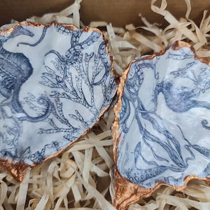 May include: Two white ceramic seashell-shaped dishes with gold trim. Each dish features a blue and white illustration of a seahorse, seaweed, and an octopus.