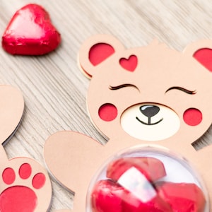 May include: Two brown cardboard bear-shaped boxes with red accents. Each box has a clear plastic dome on the front, filled with red foil-wrapped chocolate hearts. One box has a red paw print on the bottom.