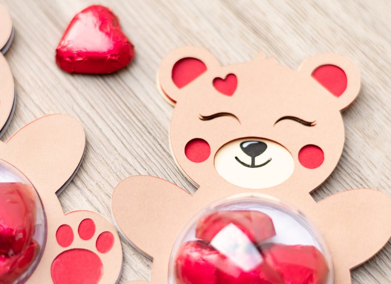May include: Two brown cardboard bear-shaped boxes with red accents. Each box has a clear plastic dome on the front, filled with red foil-wrapped chocolate hearts. One box has a red paw print on the bottom.