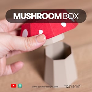 May include: A red and white mushroom-shaped box with the text "MUSHROOM BOX" on top. The box is open, revealing a beige interior. The design is geometric and appears to be made of paper or cardstock.
