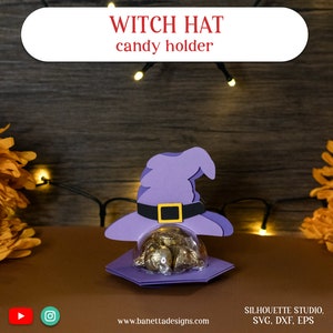 May include: Purple witch hat candy holder with a clear dome for holding treats. The hat has a black band with a gold buckle. The holder is on a wooden surface with fairy lights.
