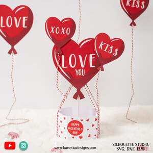 Op de afbeelding: Rode papieren hartvormige ballonnen met de woorden "LOVE YOU", "XOXO" en "KISS" zijn aan elkaar gebonden en worden vastgehouden in een witte papieren cilinder met rode harten en de tekst "HAPPY VALENTINE'S DAY".