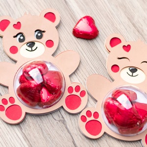 May include: Two brown teddy bear shaped cardboard boxes with red accents. Each box has a clear plastic window in the center, filled with red chocolate hearts. One box has a single red chocolate heart on the top.