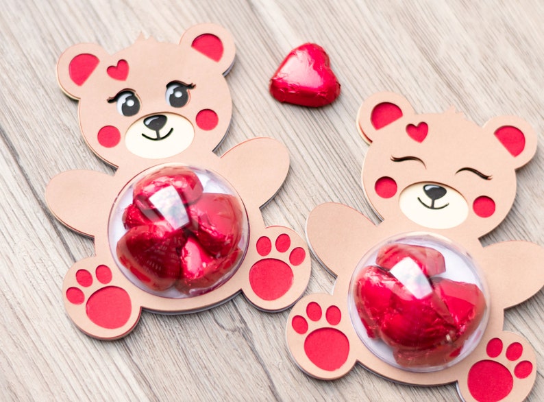 May include: Two brown teddy bear shaped cardboard boxes with red accents. Each box has a clear plastic window in the center, filled with red chocolate hearts. One box has a single red chocolate heart on the top.