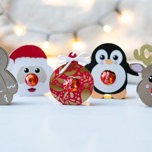 May include: Five Christmas-themed paper treat holders, each featuring a different character: gingerbread man, Santa Claus, ornament, penguin, and reindeer. Each holder has a hole in the center to hold a round chocolate candy.