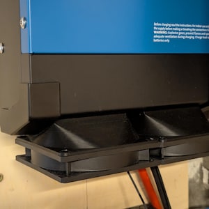 Könnte beinhalten: Nahaufnahme einer schwarzen und blauen elektrischen Komponente. Die Komponente hat zwei schwarze Lüfter und eine blaue Oberseite mit weißem Text. Der Text enthält eine Warnung vor explosiven Gasen und dem Laden von Batterien.