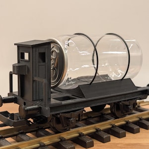 May include: A black, miniature train car with a clear, cylindrical container secured on top. The container holds a silver can. The train car sits on a section of model train tracks. The train car has a small cabin with windows.