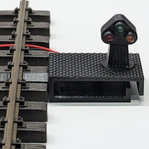 Könnte beinhalten: Ein schwarzes Modelleisenbahn-Signal mit roten, grünen und gelben Lichtern. Das Signal ist auf einer schwarzen Plattform neben einem Abschnitt von Modelleisenbahngleisen montiert. Ein rotes Kabel verbindet das Signal mit den Gleisen.