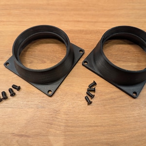 May include: Two black, square-based circular adapters with a textured finish, each featuring four corner screw holes. Accompanying the adapters are eight small black metal screws, arranged in two groups of four, intended for assembly and mounting.