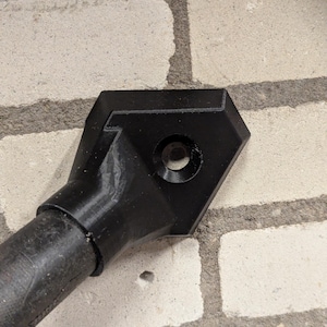 May include: A black, geometric object with a circular hole, connected to a cylindrical handle. The item is set against a textured, light-coloured brick wall. The design appears to be 3D-printed.