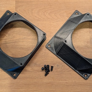 Könnte beinhalten: Zwei schwarze quadratische Lüfterhalterungen mit einer kreisförmigen Öffnung und einem kleinen Haufen schwarzer Schrauben. Die Halterungen haben ein geometrisches Design und bestehen aus einem dunklen Material. Die Artikel befinden sich auf einer hellbraunen Holzoberfläche.