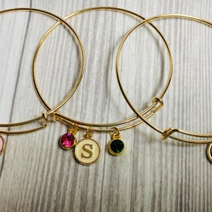 Puede incluir: Tres pulseras de brazalete doradas con dijes. Los dijes son redondos y tienen un fondo blanco con una letra dorada en cada uno. Las letras son S, S y H. Cada dije también tiene un pequeño cristal unido a él. Los cristales son verde, rosa y transparente.