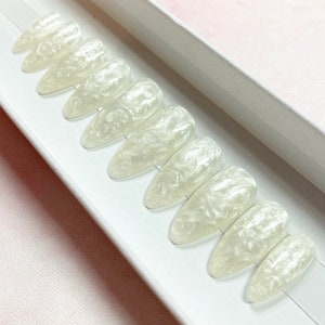 Op de afbeelding: Een set van tien parelmoer, amandelvormige valse nagels in een witte doos. De nagels hebben een wervelend, gemarmerd effect in wit en crème, waardoor een lichtgevende uitstraling ontstaat. De nagels zijn een schoonheidsproduct.