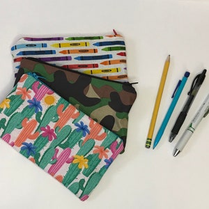 Puede incluir: Tres estuches de lápices de tela con diferentes patrones. El estuche superior tiene un patrón de crayones coloridos, el estuche del medio tiene un patrón de camuflaje y el estuche inferior tiene un patrón de cactus.