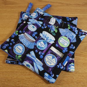 May include: Two black fabric pot holders with blue trim. The pot holders feature a repeating pattern of blueberry jam jars with blue and white labels. The labels say "Homemade Blueberry Jam" and "Blueberry Jelly".