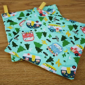 May include: Two square pot holders with a light teal background. The fabric features a repeating pattern of colorful camper vans, green trees, cacti, and crescent moons. Each pot holder has a yellow loop for hanging.