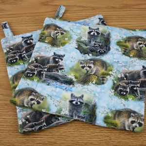 May include: Two square potholders with a light blue background featuring a repeating pattern of raccoons, trees, and white flowers. Each potholder has a small fabric loop for hanging. The potholders are made of quilted fabric.
