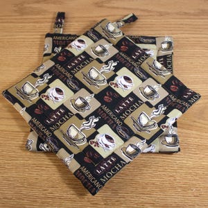 May include: Two black and brown fabric pot holders with a checkered pattern and coffee themed text. The text includes "Latte", "Mocha", and "Americano".