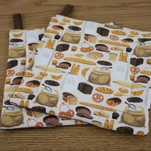 May include: Two square pot holders with a white background and a repeating pattern of bread, rolls, and flour sacks. Each pot holder has a brown fabric loop for hanging. The words "Fresh Bread" are printed on the pot holders.