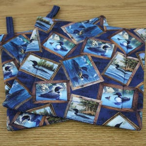 May include: Two blue potholders with a repeating pattern of framed images of loons on a lake. The potholders have a dark blue background and a loop for hanging. The frames are brown and the loons are black and white.