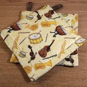 May include: A pair of yellow pot holders with a musical instrument pattern. The pot holders feature a variety of instruments, including a violin, a trombone, a french horn, a tuba, a drum, and a cello.