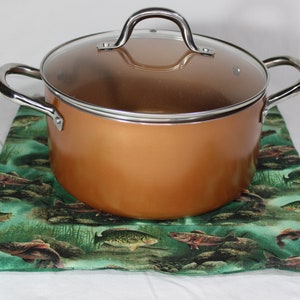 May include: A copper pot with a glass lid sits on a green and brown pot holder with a fish pattern.