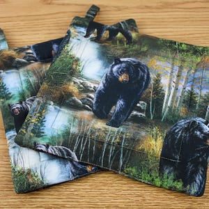 May include: Two square pot holders with a loop for hanging. The pot holders feature a detailed print of black bears in a forest setting with a river. The background includes trees, rocks, and foliage. The pot holders are quilted.