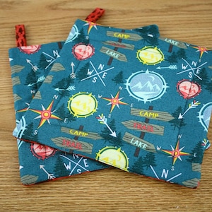 May include: Two teal pot holders with a camping theme. The fabric features compasses, arrows, trees, and signs that say "Camp" and "Lake". The edges are trimmed in red, and each has a red loop for hanging.