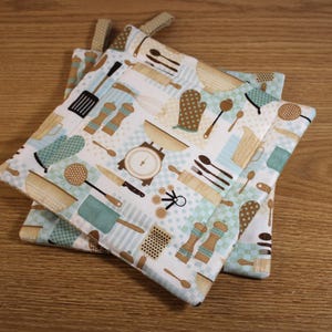 May include: Two blue and white kitchen pot holders with a brown and white pattern of kitchen utensils and spices. The pot holders are made of fabric and have a quilted texture.