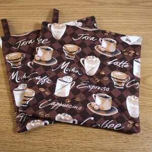 May include: Two brown and white cotton pot holders with a coffee theme. The pot holders feature various coffee drinks, including cappuccino, espresso, latte, mocha, and java. The pot holders have a checkered pattern and the words "Coffee", "Java", "Mocha", "Cappuccino", "Espresso", and "Latte" are printed on them.