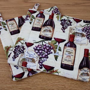May include: Two quilted pot holders with a wine theme. The cream-colored fabric features images of wine bottles, glasses, and purple grapes. Each pot holder has a loop for hanging.