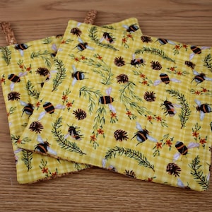May include: Two yellow pot holders with a bee and floral pattern. The pot holders have a gingham pattern and are decorated with bees, pinecones, and sprigs. Each pot holder has a brown loop for hanging.