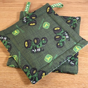May include: Two green and black fabric pot holders with a repeating pattern of John Deere tractors and logos. The pot holders are made of a textured fabric and have a white border.