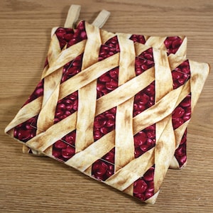May include: Two cherry pie patterned pot holders with a woven design. The pot holders are made of fabric and have a loop for hanging.