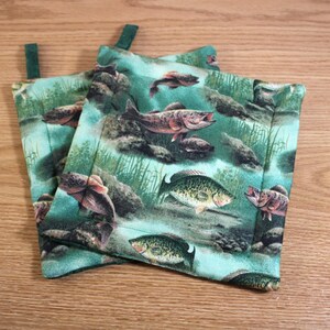 May include: Two green and blue pot holders with a fish print. The pot holders have a loop for hanging.