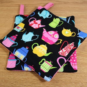 May include: Two black fabric pot holders with a tea pot pattern. The pot holders have pink and white polka dot fabric on the back. The pot holders have pink, yellow, blue, green, and orange teapots with a variety of designs.
