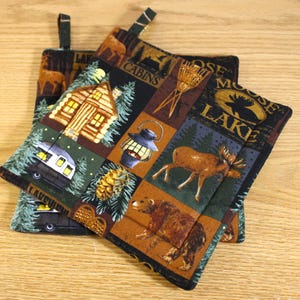 May include: Two black and brown quilted pot holders with a woodland theme. The pot holders feature a cabin, a moose, a bear, a lantern, a pine cone, and the words "Moose Lake Cabins".