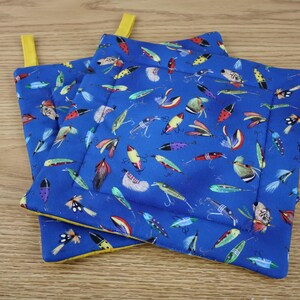 May include: Two blue square pot holders with a colorful fishing lure pattern. The pot holders have yellow trim and a yellow hanging loop. The pot holders are made of fabric and are designed to protect hands from heat.
