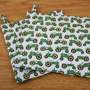 May include: Two white square pot holders with a green tractor pattern and yellow wheels. Each pot holder has a small fabric loop for hanging. The pot holders are made of fabric and are on a wooden surface.