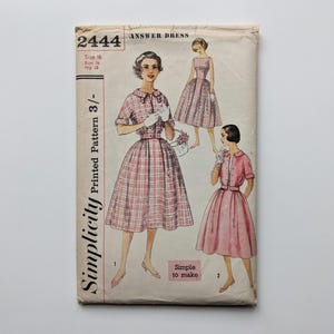 Könnte beinhalten: Vintage Simplicity-Schnittmusterumschlag mit Kleiderillustrationen. Der cremefarbene Umschlag hat oben die Nummer 2444. Die Kleider sind in rosa und rot kariert und in einfarbigem Rosa. Der Text "Answer Dress" steht oben.
