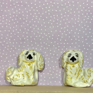 Könnte beinhalten: Zwei kleine weiße Keramikfiguren von Hunden mit goldenen Akzenten. Die Hunde sitzen mit geneigtem Kopf und Blick nach vorne. Die Figuren stehen auf einem hellvioletten Hintergrund mit weißen Punkten.