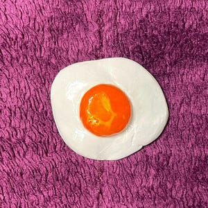 May include: A white ceramic egg with a bright orange yolk. The egg is sitting on a purple textured surface.