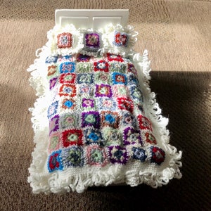 May include: A white doll bed with a crocheted patchwork quilt and two pillows. The quilt is made of colorful squares in shades of red, blue, green, purple, and white.
