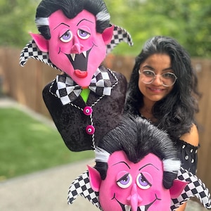 May include: Two plush Count Dracula character decorations. The pink-faced figures have black hair, white streaks, and checkered collars. One is attached to a black jacket with pink buttons. The other is held by a person.