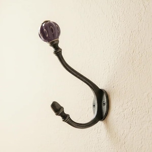 Peut inclure: Un crochet porte-manteau mural en métal avec une finition sombre et patinée. Le crochet présente un design incurvé à deux branches, et la branche supérieure est surmontée d'un bouton en céramique violet foncé, nervuré et brillant.