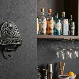 May include: A wall-mounted, cast iron bottle opener with a dark grey finish. The opener features a relief design of a tree above the embossed text "GIN & TONIC". It is mounted on a dark grey wall with a subtle, repeating fan-shaped pattern.