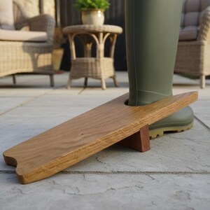 Op de afbeelding: Een houten laarzenknecht met een gladde, lichtbruine afwerking en een centrale ronde uitsparing, voorzien van een klein steunblok aan de onderzijde. Het voorwerp staat op een stenen terras, met een hoge olijfgroene rubberen laars in de uitsparing.