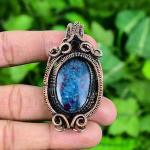 Peut inclure: Un pendentif en fil de cuivre enveloppé avec une pierre précieuse bleue et rouge. Le pendentif a un design détaillé avec un travail de fil complexe.